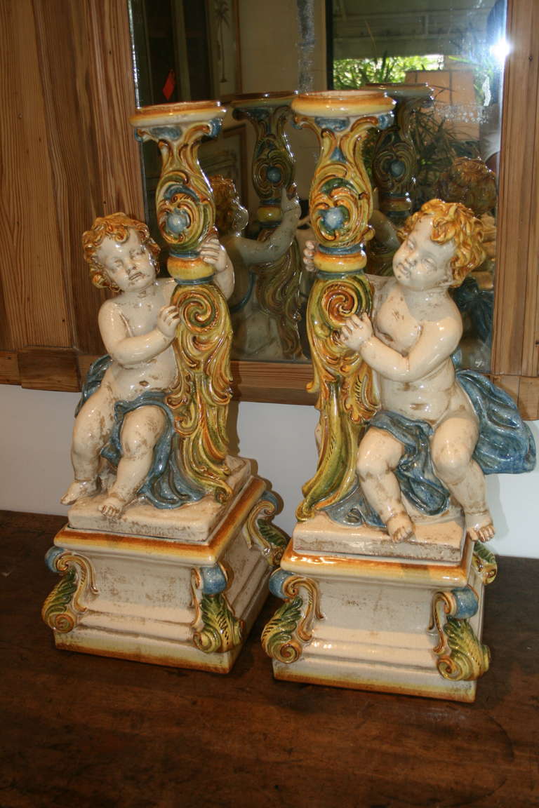 Italian Majolica of golden haired putti, set on rocks beside and embracing acanthus detailed candleholder, resting on separate scroll cornered square base; both pieces of glazed terracotta with cream, blue, green and gold finished details.