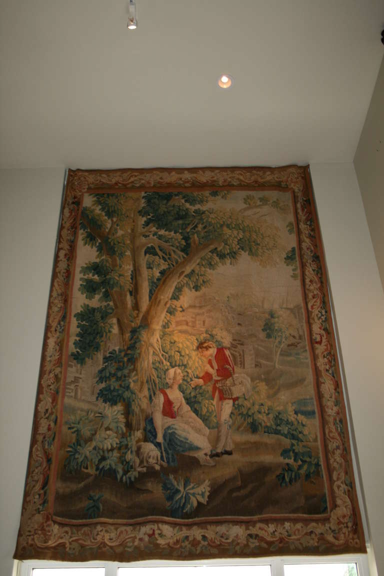 Very charming 18th century tapestry with young man and maiden conversing under a tree with dog, a village in the distant background and framed with a  border of floral and leaf designs with protective back fabric panel.