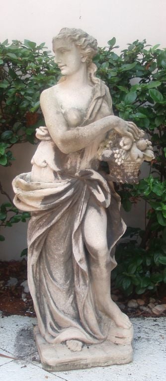 Well detailed cast Vicenza stone garden statue of a young lady standing in robes and holding a basket of fruit, head slightly turned to the right; naturally weathered finish.