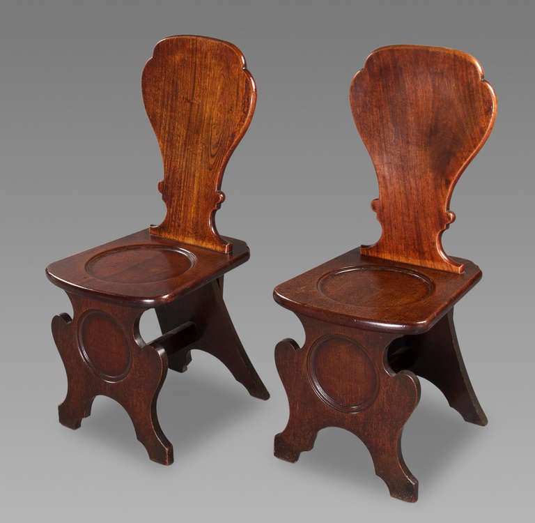 A good pair of George III mahogany hall chairs, each with a slightly dished waisted back, dish seat, and raised on scrolling front support, united by a stretcher to the rear. Examples of these chairs were produced in English furniture from at least