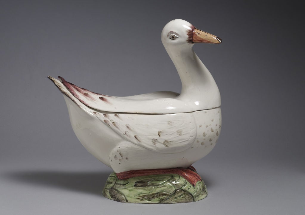 An amusing and rare Faience Goose tureen and cover, the seated bird bends its smiling head to one side, forming the handle, its upper body forming the cover and the lower body forming the base, resting on a green oval mound.