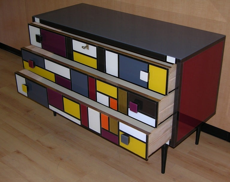 a very chic commission-made chest with 3 drawers, surrounded with colored glass, decorated with a geometrical pattern inspired by Mondrian's work, quality details of manufacturing with glass covered slanted handles, on black lacquered legs, with