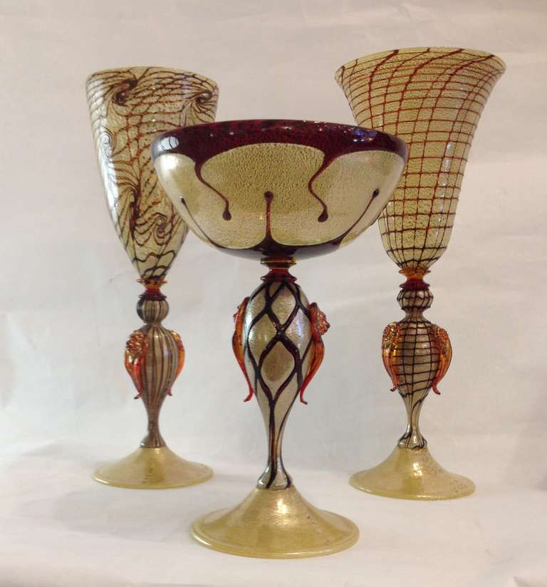 A set of three vintage 1970s gold and red Murano glass hand crafted vessels raised on foot. Each piece is individually blown and detailed with a different free hand applied deep rich red overlay. The glass bodies are worked with expanded pure gold