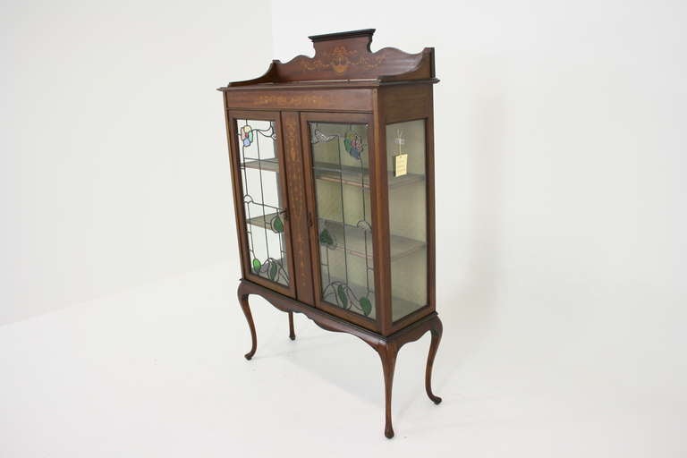 A beautiful Art Nouveau mahogany inlaid display cabinet with inlaid ledgeback  top above a pair of stained glass leaded doors enclosing a green velvet-lined interior with two shelves ending on tall cabriole legs.