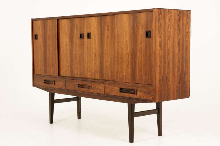 Nice rosewood sideboard from Denmark dating to the 60”²s. Tall sideboard shows four sliding doors which open to a mirrored back with a glass shelf and three drawers, on the left. The middle and right doors opens to the two adjustable shelves and