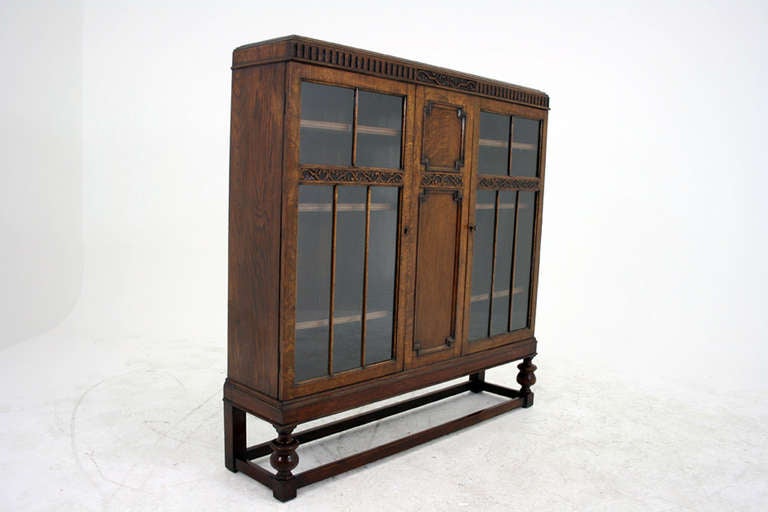 Solid oak bookcase, display cabinet with rectangular top, carved frieze, carved blank door, flanked by two astragal glass doors, enclosing three adjustable wooden shelves, ending on turned bulbous feet connected by stretchers.  Excellent original