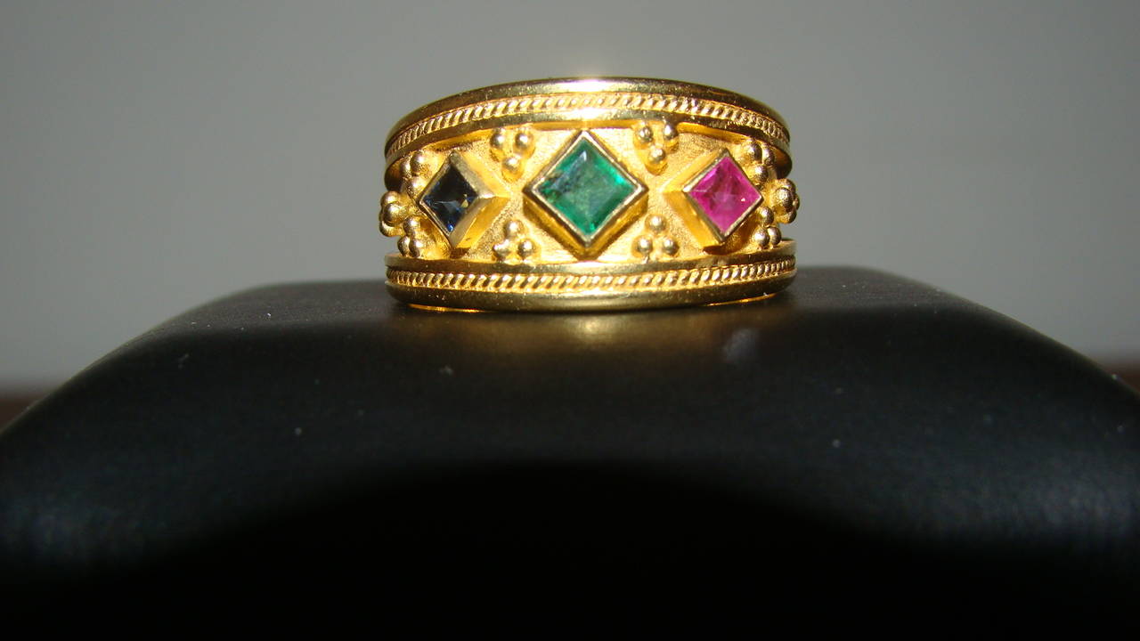 This beautiful Etruscan ring features a kite set emerald in the center with a kite set sapphire and ruby on either side. Size 8.5. Face measures about 11.5 mm tall and 21 mm wide. Weighs 8.7 g. Fully hallmarked 750.