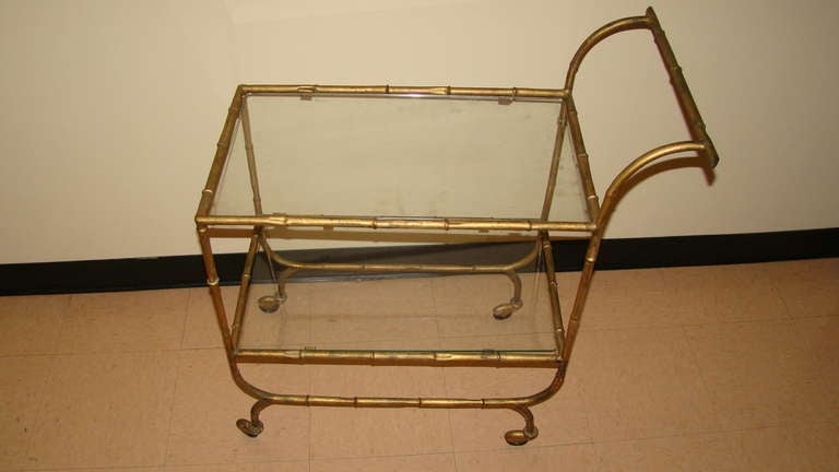 Outstanding Faux Bamboo Gold Guilded Metal Rolling Bar Cart. This beautiful piece is comprised of faux bamboo metal with gold guild finish. It features two tiers with glass and wheels. Perfect for serving.