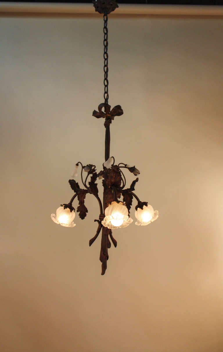 Handcrafted in France during the first decade of the 20th century, this gorgeous chandelier features blown satin glass shades in a tulip style. Beautiful, organically-inspired bronze detail work can be found throughout the chandelier, characteristic