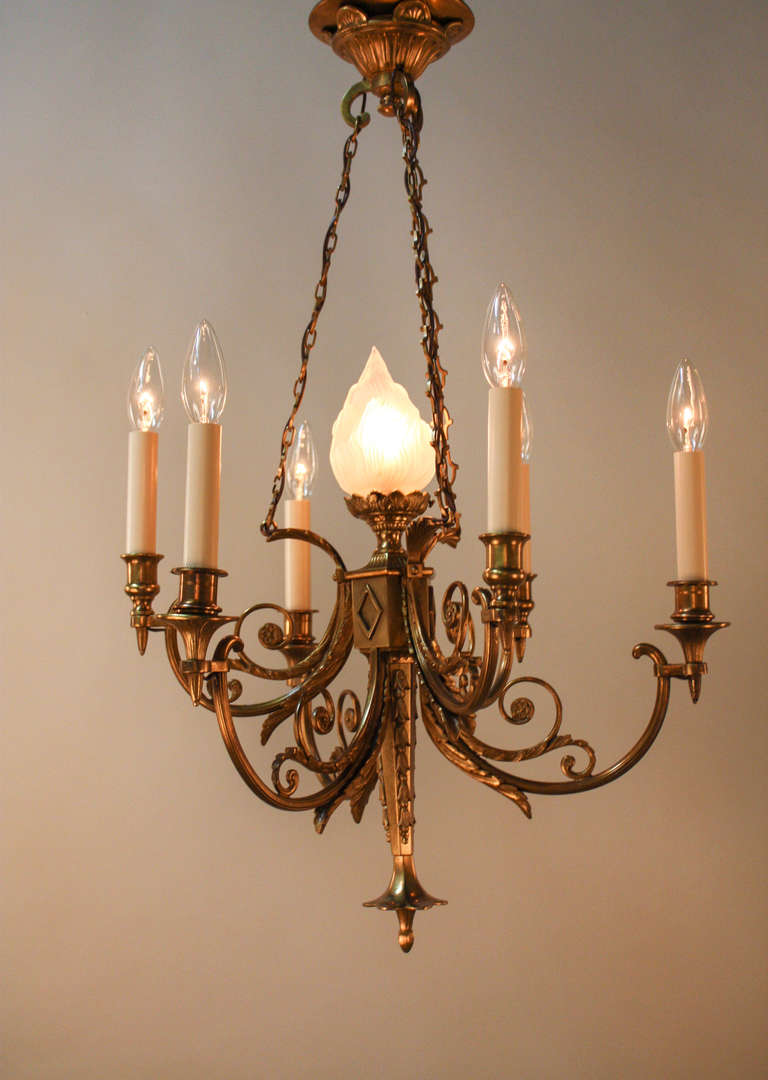 Crafted in France during the 1930s, this stunning chandelier features seven lights and the fantastically detailed bronze work. 

The bronze body features curling arms which stretch from each of the lights to the glass shade torch in the center of