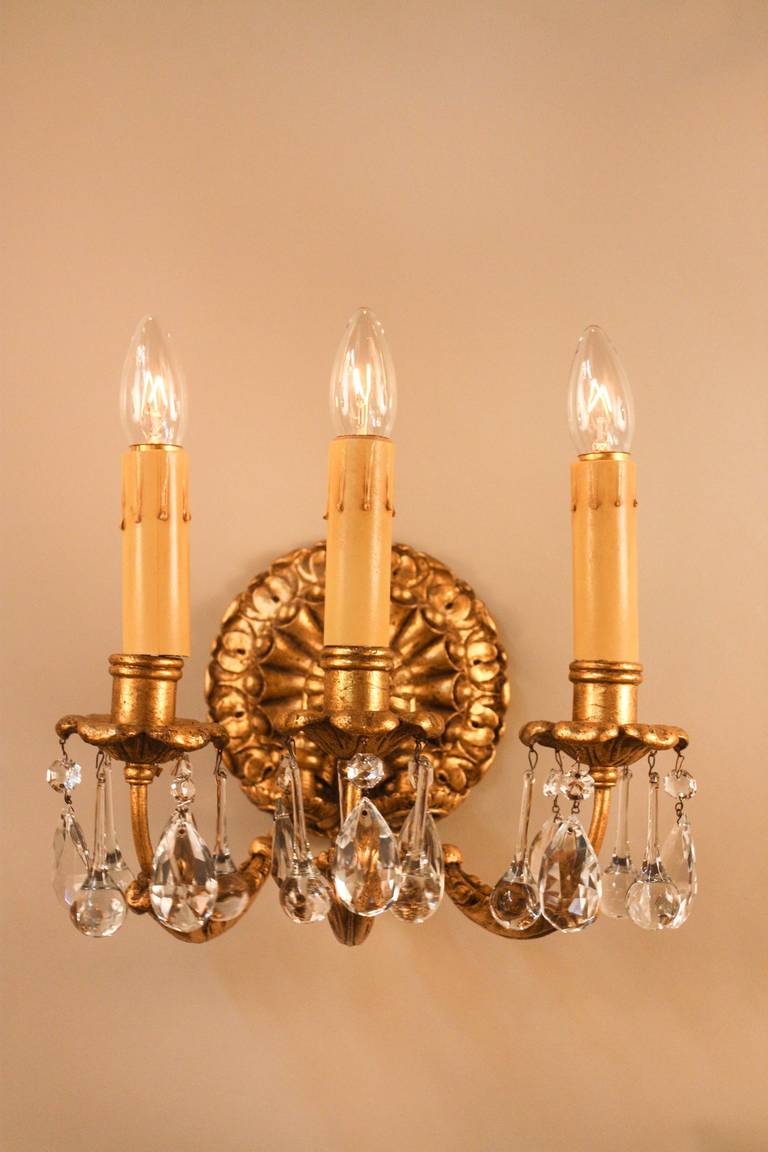 Ursprünglich wurden diese Wandleuchten in den 1950er Jahren in Italien hergestellt und über die Sterling Bronze Co. in New York importiert und verkauft. Die dreiflammigen Wandleuchter sind mit eleganten:: hängenden Kristallen und Blattgold auf Holz