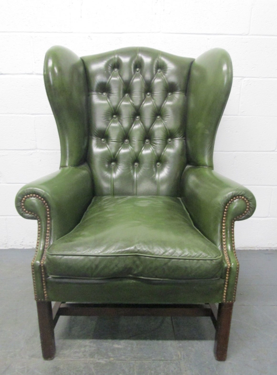 Green leather tufted wingback chair. Has a walnut base, loose cushioned down seat and brass studded trim.