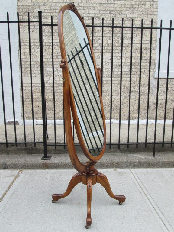 Oval shaped standing mirror on a pedestal base with pad feet and wheels. Wood has some carvings to the top and bottom.