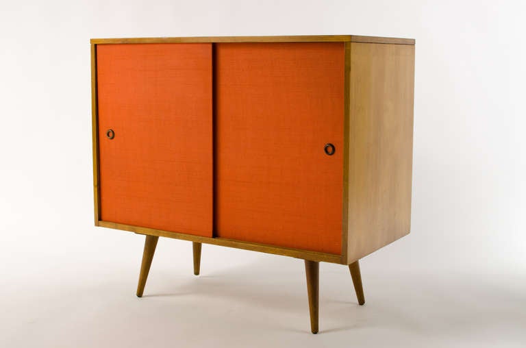 Exceptional small credenza with rare orange grasscloth sliding doors designed by Paul McCobb. Removable and adjustable shelfs on each side. Doors are a greenish brown color. Stunning piece. All original and signed.