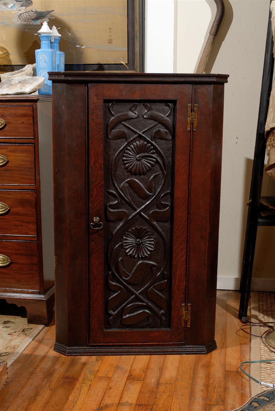 19th Century English hanging corner cabinet of oak with a molded crown above an inset door with a ribbon and floral carved panel hinged with brass hardware and flanked by canted corners and having molding along the base as well.  The cabinet is
