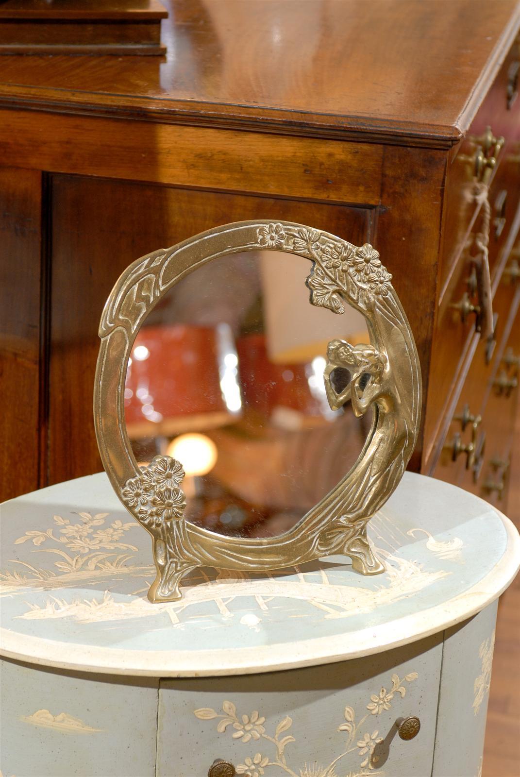 An Art Nouveau bronze, easel-back frame, dressing mirror w/full figure of woman in a floral motif.  The front of the woman's face can be seen in the mirror's reflection.