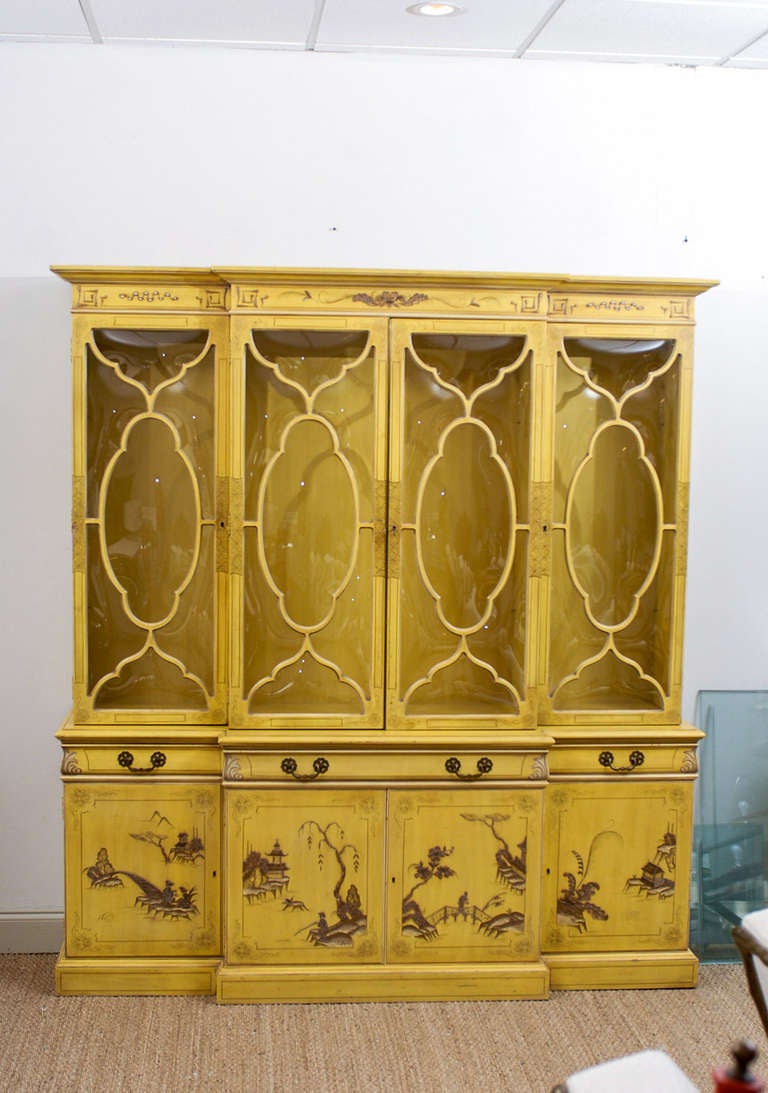 The Chinoiserie Breakfront Bookcase in canary yellow has a decorative painted crown above four curved glass doors that open to adjustable glass shelves on the interior. The center drawer is fitted with a leather top desk and flanked by drawers on