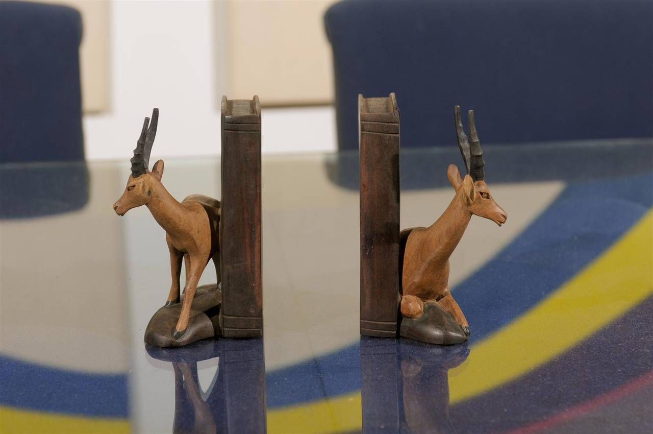 Friendly pair of vintage hand-carved bookends made of wood and having three different wood finishes. Each has a stylized carved gazelle, one seated and one standing, on top of a carved rock and resting against a carved book.