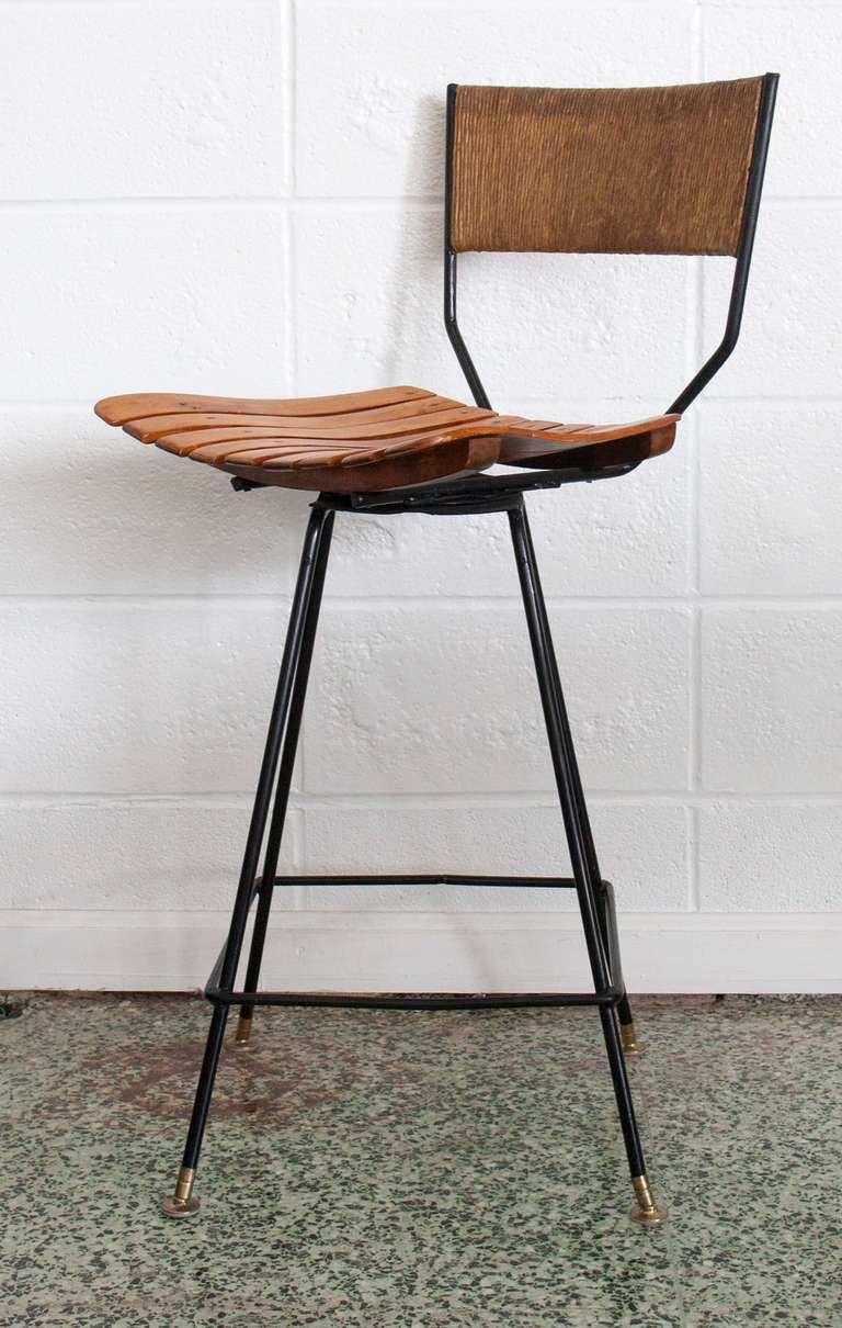 Circa 1958 set of four swivel bar stools designed by Arthur Umanoff for Raymor. Swivel stool seats are bar height at 28.75” high. Narrow black wrought iron base with foot rest, slat wood seats and woven paper rope backrests. 

Seat Height: