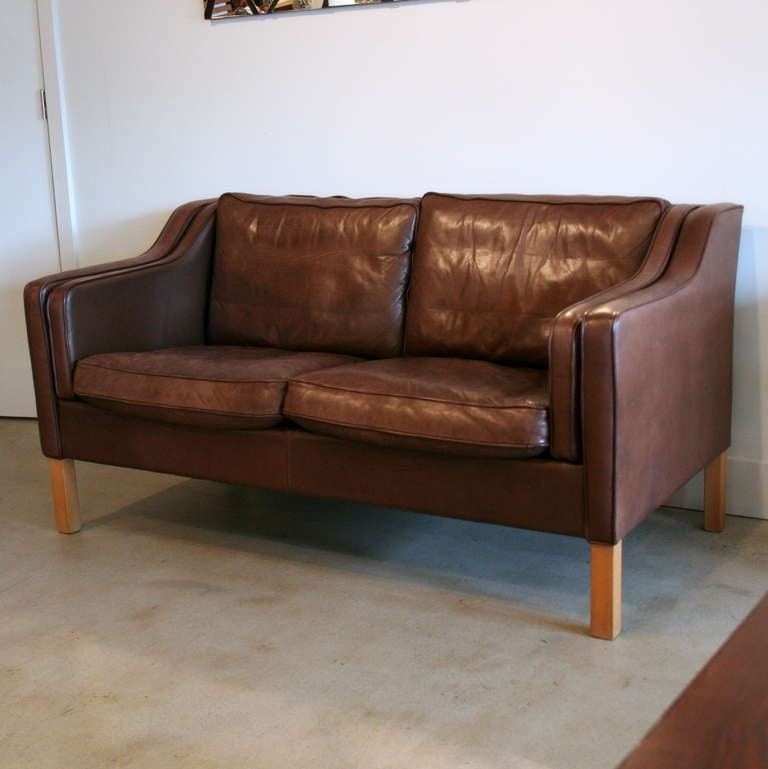 A perfectly distressed 2-seat leather sofa made in the classic mid-century style of Borge Mogensen. With a warm medium brown patina on the leather, down-wrapped cushions with piping detail, and light wood legs, this sofa provides plenty of welcoming