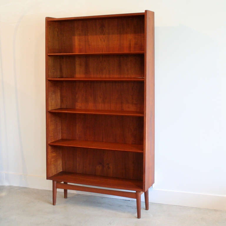 Tall teak bookcase with gently angled front profile. Adjustable shelves feature beveled edges, and it all sits on solid teak legs, raising it off the ground for a more elegant aesthetic.