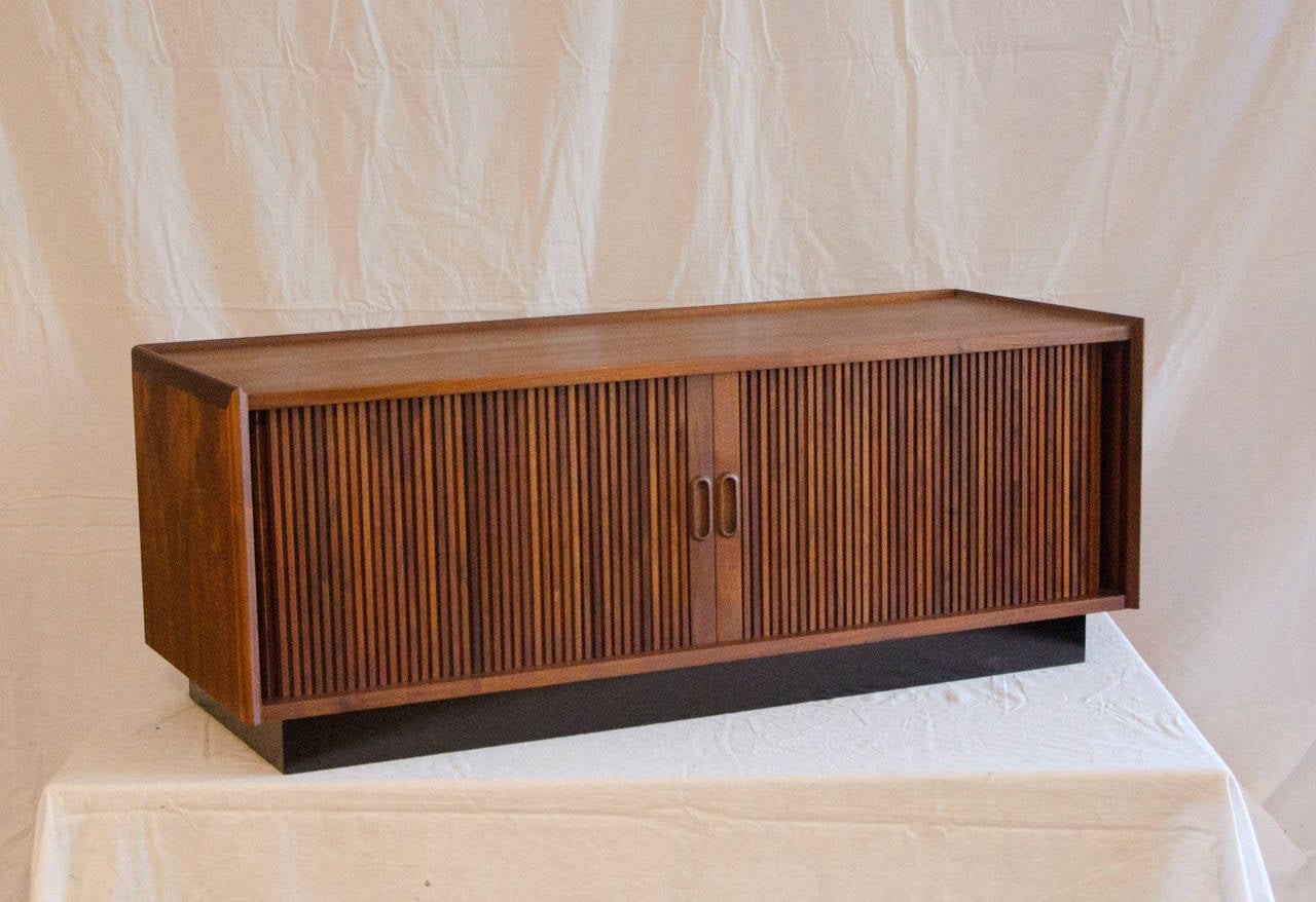 Medium size walnut cabinet with tambour doors. Cabinet has two dividers which are removable. A black plinth base supports the walnut cabinet. The top surface has a 1/2