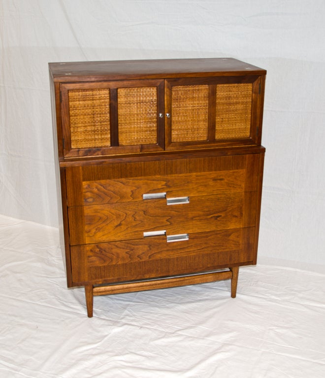 Mid century walnut hiboy by American of Martinsville, three large drawers with asymmetrical aluminum handles in lower section, upper section of chest has two interior drawers behind cane front doors. Aluminum inlays on top surface. Retains the