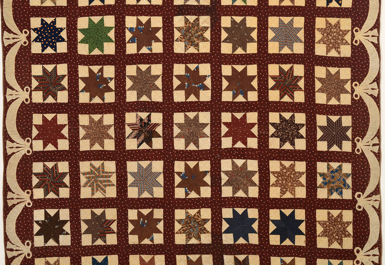 This Lemoyne stars quilt is made of a beautiful variety of fabrics and unusual color choices. The background and swag and bow border are a pale marigold colored print that is beautifully offset with the brown grid. Condition is very good but not