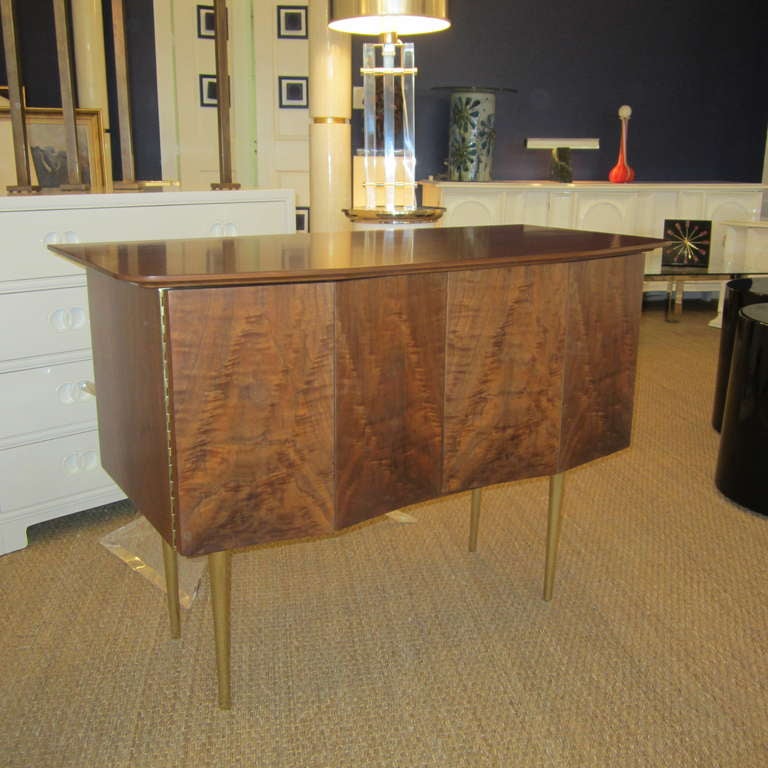Cabinet fashioned of burl wood in front and walnut on top and sides. The front is angled and the doors are bifold; they reveal a shelf on one side and two drawers on the left. The legs are tapered polished brass and the piano hinges follow suit in