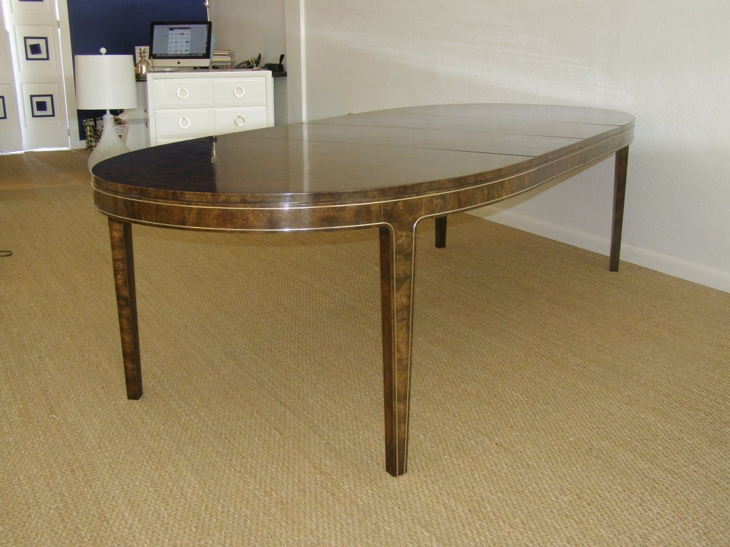 Exquisite dining table fashioned in burled amboyna wood with brass racetrack fenestration. With gently tapered legs, this table comes complete with two extension leaves, shown, which feature the finished apron with brass fenestration. 
Full set of