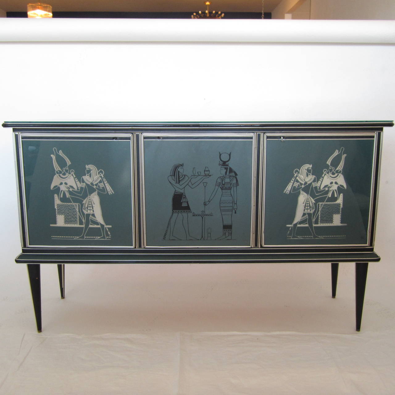 Great Mid-Century Italian credenza with tapered legs and three glass front doors with Egyptian reverse painted motif. The body is in green pebbled vinyl over wood with black vinyl and aluminum trim. The doors have the original skeleton key with