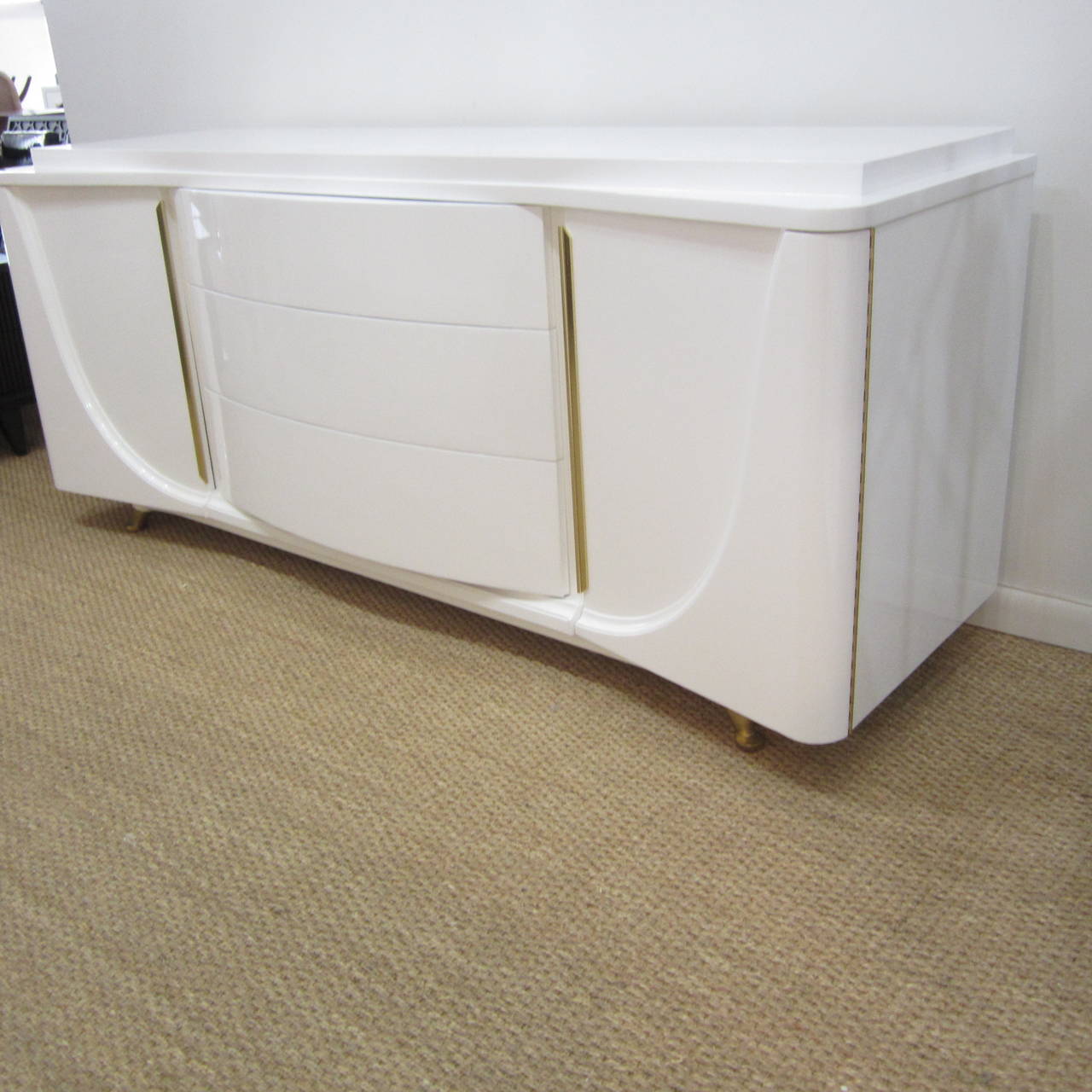 Beautiful low sitting credenza superbly finished inside and out in high gloss white lacquer with vertical brass door pulls. The top has a flat surface on top of a sweeping, curved second tier that flows with the curvaceous lines of the body and