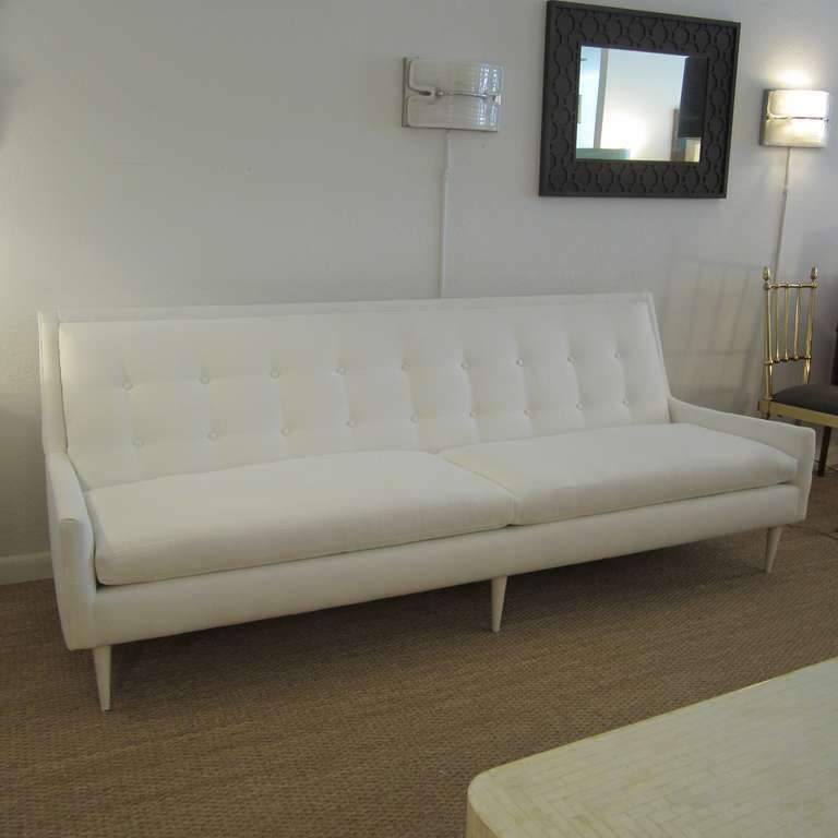 Elongated sofa with a slight outward flare to the arms. Framing is comprised of six tapered legs that are white lacquered which show some signs of usage. Back has double row of buttons while seat is two long cushions. This elegant period sofa is