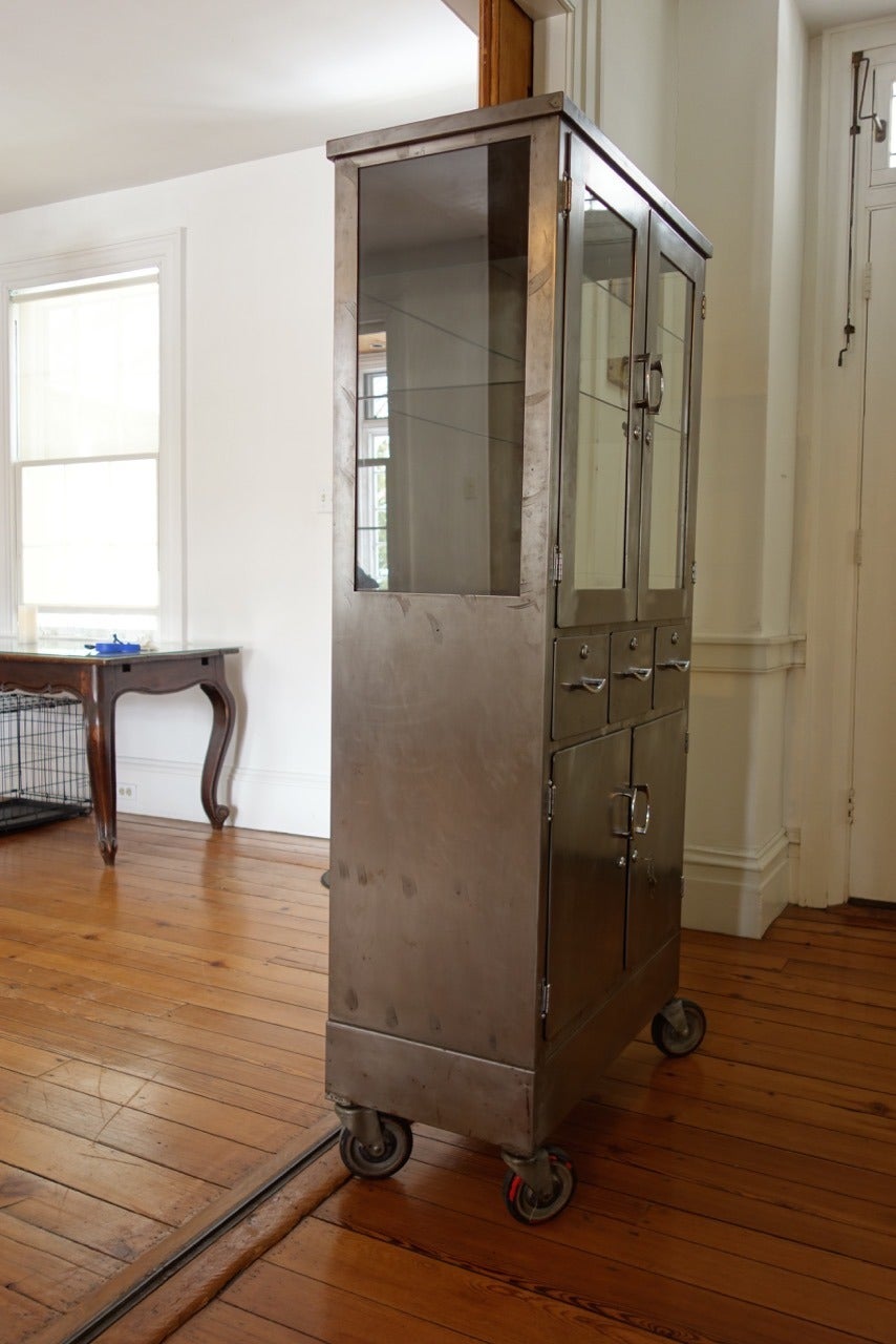 Beautiful vintage metal and glass medical cabinet on original wheels. Cabinet has glass sides and front with two glass interior shelves, three small drawers, bottom has double doors with shelf. All locks and keys. Some age appropriate wear but