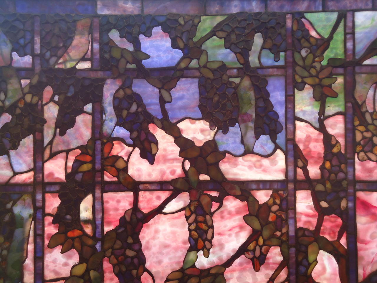 Intricate stained glass window with grape motif, unusual pink glass with greens and blues, in the style of Louis Comfort Tiffany, mid-20th century.
Grapes are raised from the background glass in a textural masterpiece.