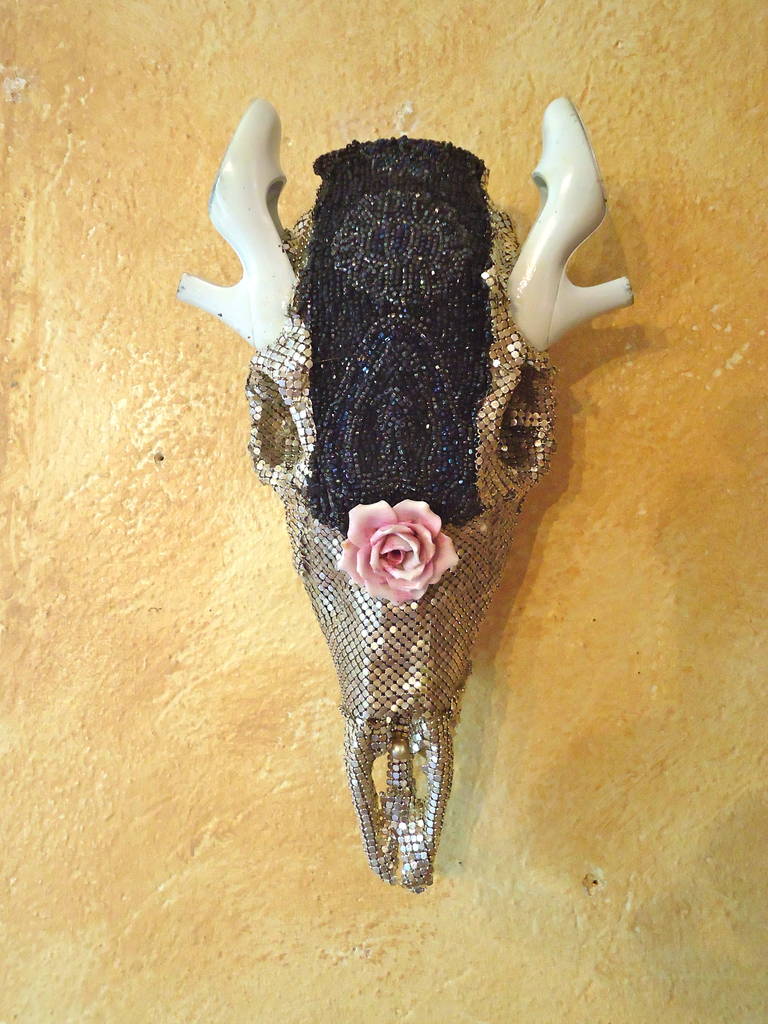 Small skull transformed into gorgeous wall art sculpture and mix-media ambrosia by artist Fay Sciarra. Encrusted in bits of antique silver mesh from old pocket books, a navy beaded purse remnant, a pink porcelain rose and white lacquer high heel