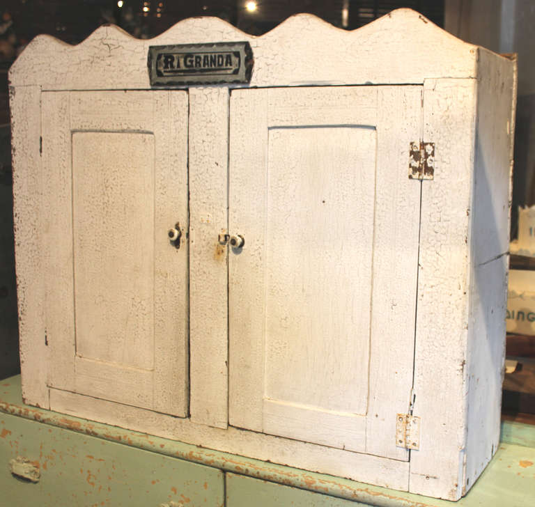 Folky American Painted hanging cabinet. White crackle, alligator paint on exterior of this 2 door cabinet. The scalloped top has a mirrored name plate, R. Granda. The interior doors a painted charcoal and have yellow chalk writings. One such has the