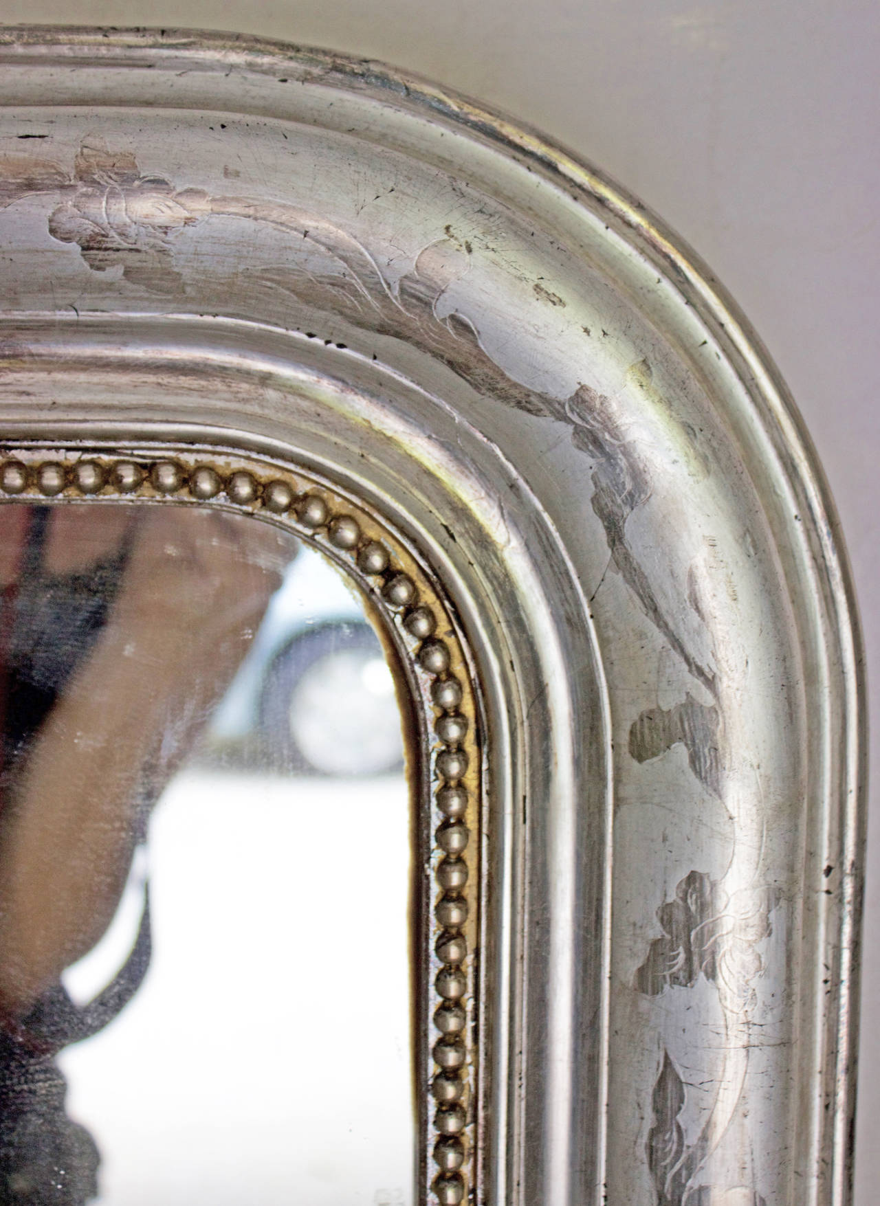 Rounded edges at the top and a row of silvered wooden pearls around three sides of the glass.This mirror has its original silver leafed gilt frame with a light and mat subtle foliage incised pattern all around except the bottom part, as it should be