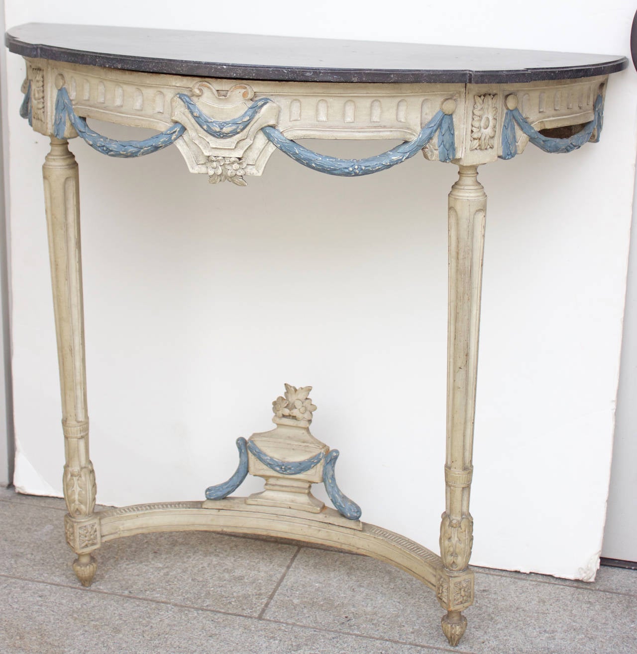 A very fine carved and painted wood console “en demilune” with its original black marble top above a frieze with hanging laurel garlands painted in blue. The console is supported by tapered fluted legs joined by a stretcher surmounted in its center