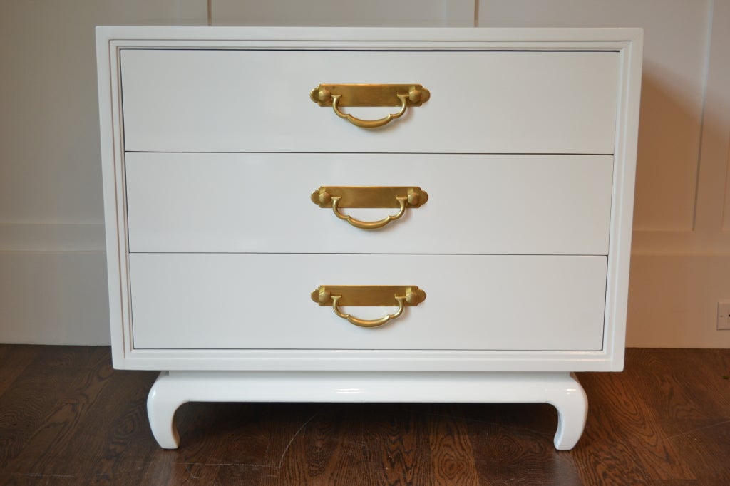 Newly lacquered in gloss white, this commode has a subtle oriental form with handsome solid brass hardware.