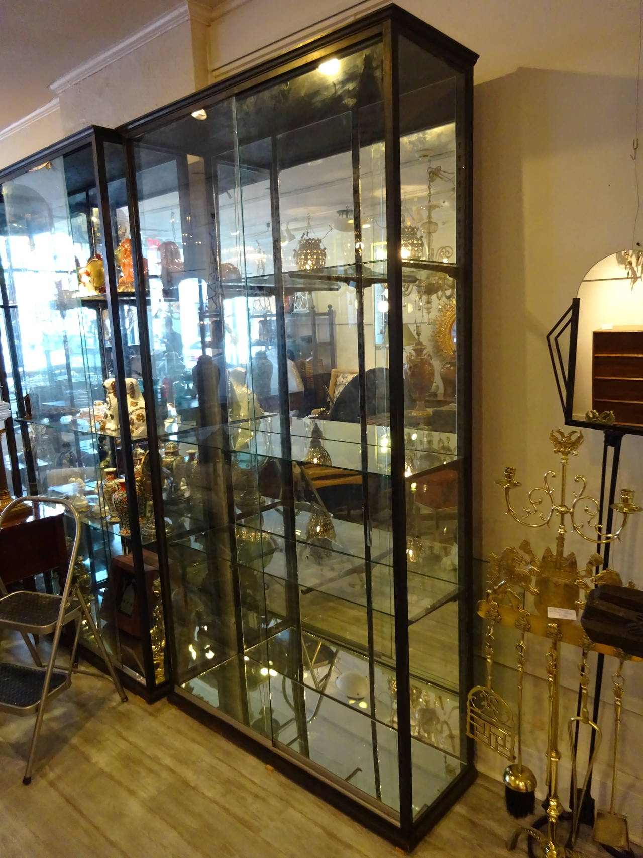 Two antique neoclassic bronze showcases each one with dark brown patina, tall and narrow rectangular form with mirrored back and two glass doors. The interior is fitted with the original bronze brackets supporting three glass shelves. A third one is