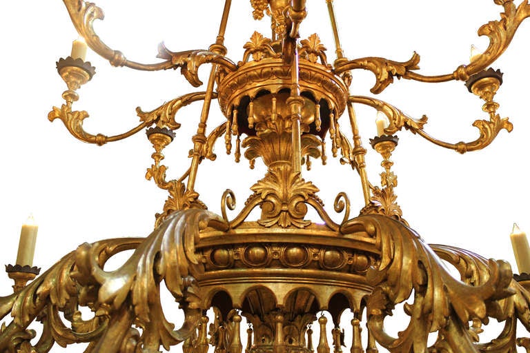 A towering 19th century tuscan giltwood palazzo chandelier, multi-tiered accented with giltwood pendants, scalloped metal bobeches and elaborately carved with acanthus leaves, egg and dart, floral and foliate motifs, centered with an urn overflowing