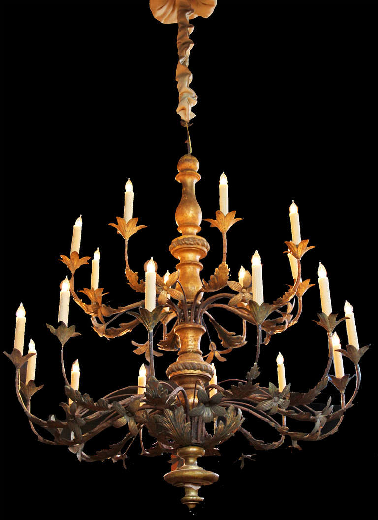 An 18th century twenty-light Italian giltwood, parcel-gilt tole and wrought iron chandelier, with two-tier of lights (now electrified) emanating from a turned giltwood standard with branch-like arms embellished with foliate and blossoming floral