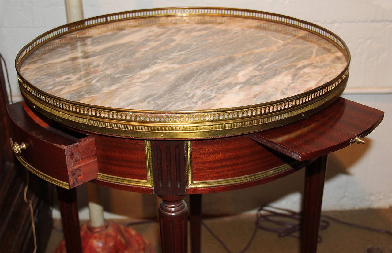 A pair of 19th century French mahogany bouillotte tables, the marble top surrounded by a pierced brass gallery (a removable felt-like round insert would have been used for bouillotte card games), above the conforming frieze, fitted with two drawers