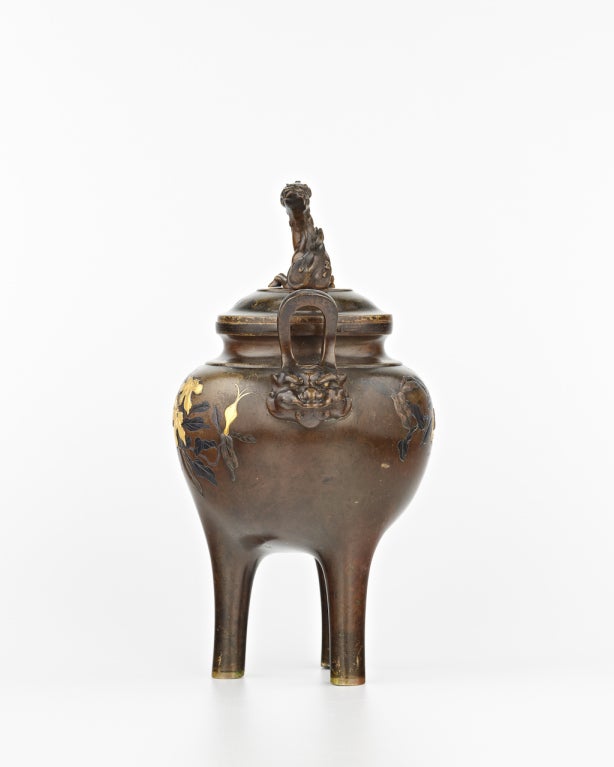 The bronze censer resting on three tall cylindrical feet and flanked with two handles modeled as beast's heads, the body inlaid with gold, copper and shakudo, one side depicting a branch of peaches, the other side with a bird and sprays of flowers,