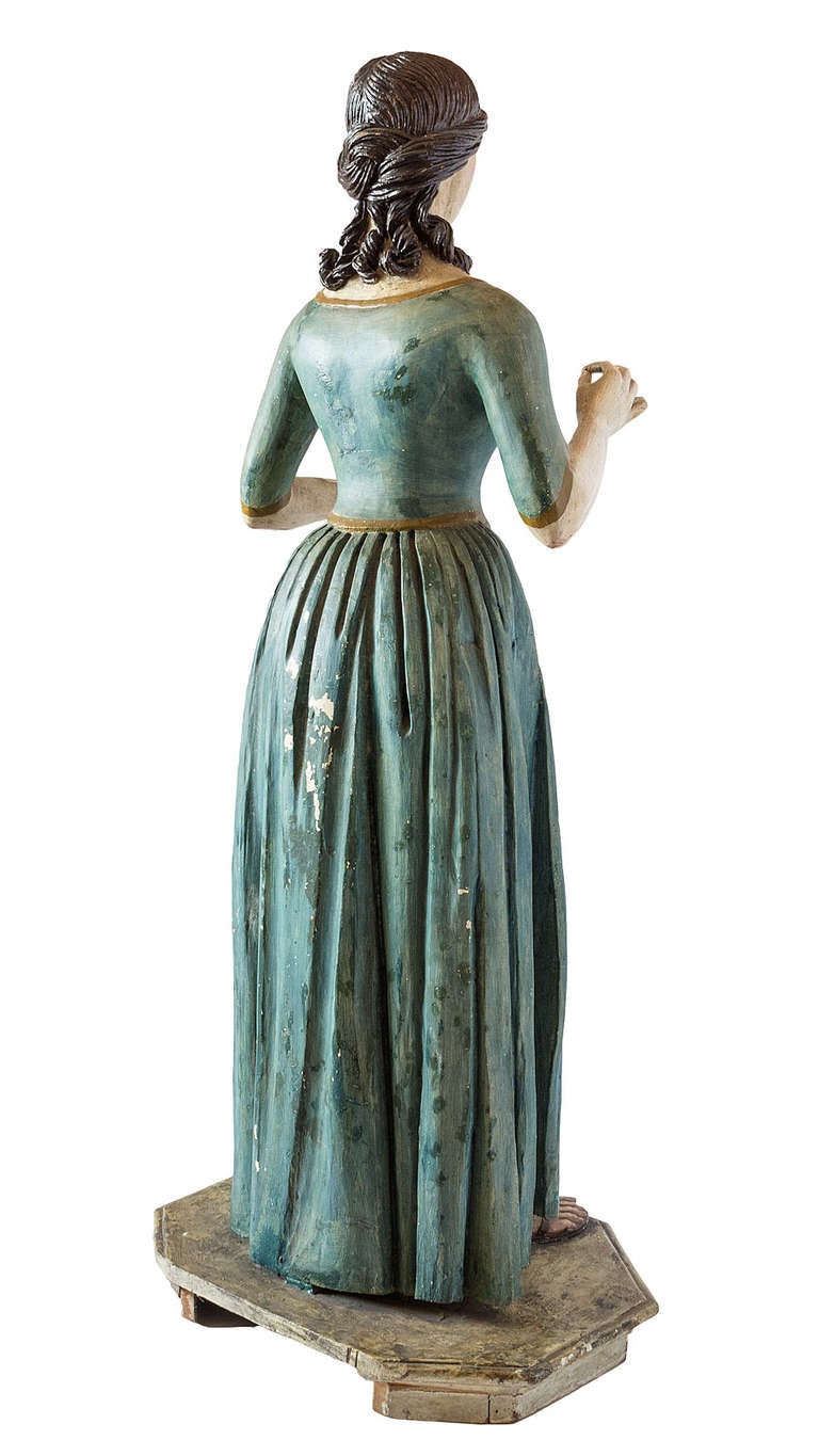 A beautifully carved wooden figure decorated with paint over gesso. The face, hair and folds of the dress are described with great attention to realism.The figure is attached to the pedestal.