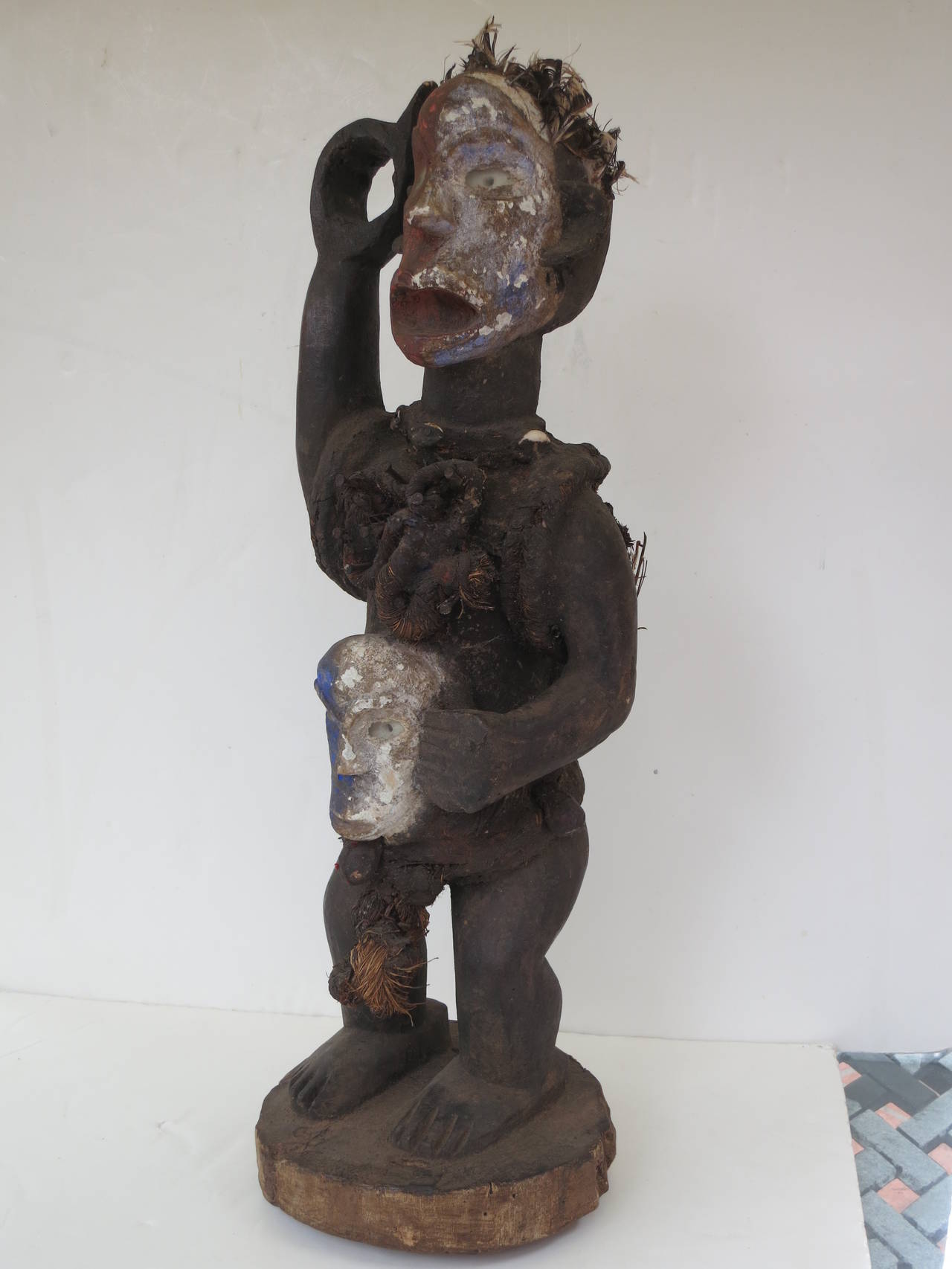 This Bakongo power figure is adorned with feathers, shells, nails and raffia. The eyes of the figure and the mask he holds are of inset glass. Magic bundles are attached to the torso both front and back. Paint is applied to the face of the figure