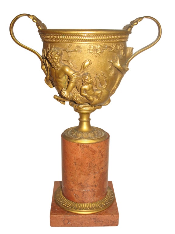 This is an antique gilt bronze urn atop a classic, cylindrical marble pedestal. Centaurs and putti encircle the cup and there is an overall fineness of sculptural details.