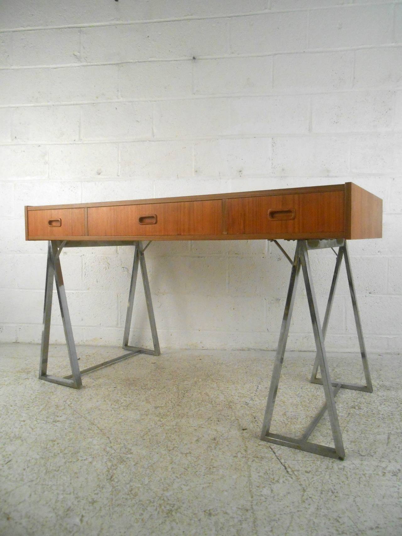 Unusual teak desk with simple modern design set on two polished chrome saw horse legs. Three drawers with sculpted handles, wide surface area with warm teak grain, 'A' frame open legs. Finished back, perfect for open floor plans.

(Please confirm
