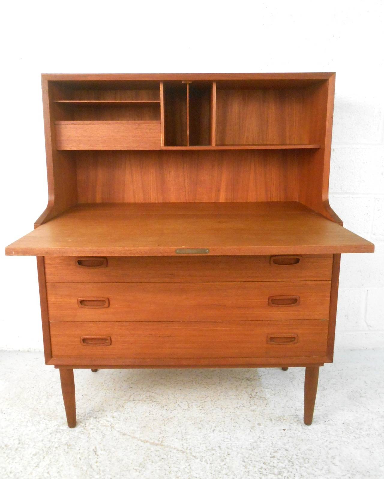 This teak drop front offers the perfect workspace for tight quarters, including three drawers for added storage and a desk top organization area. Tapered legs and carved drawer pulls add to it's Mid-Century charm. Please confirm item location (NY or